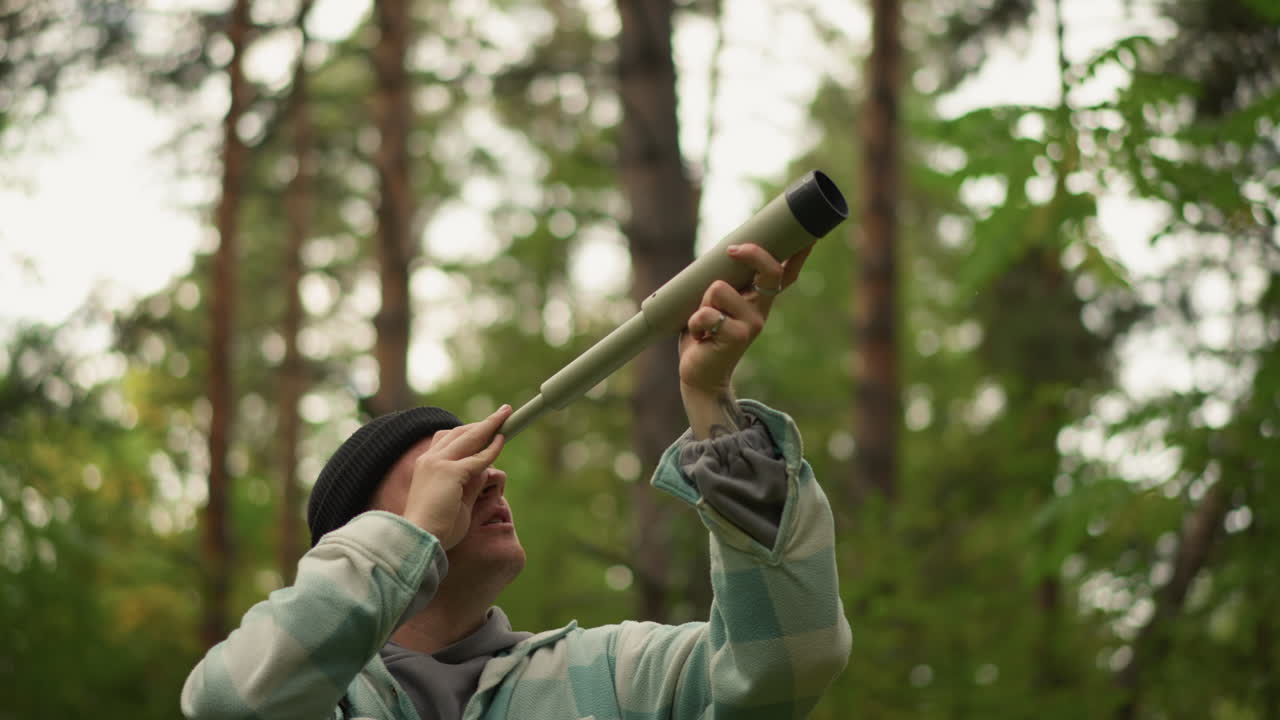 Hiker examines forest surroundings attentively, Mountain adventurer uses binoculars to scout pine canopy, Enthusiastic trekker with handmade telescope surveys majestic tall pine trees above