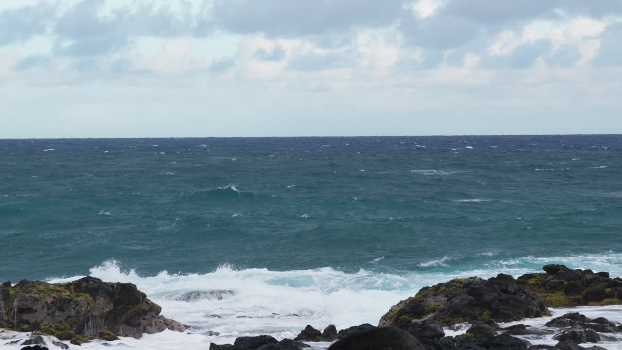 A calm yet powerful seascape featuring turquoise ocean waters extending to the horizon, with gentle rolling waves breaking against volcanic rocks in the foreground beneath a partly cloudy sky