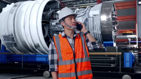 asian male engineer with safety helmet talking on smartphone while standing with airplane engine maintenance conducted, analytics checking the turbine