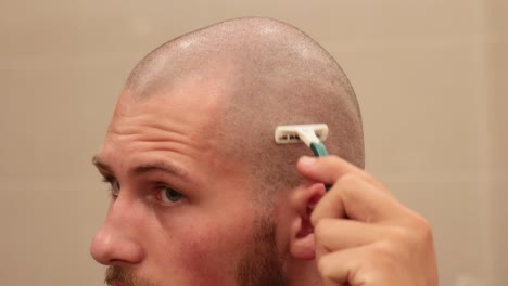 close up of young man shaving his own bald scalp with disposable razor inside bathroom