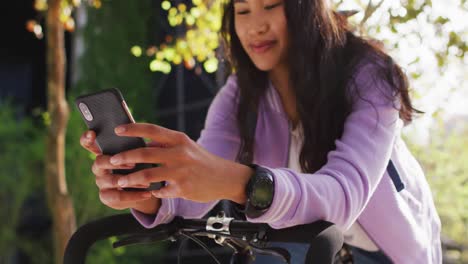 mujer asiática con bicicleta sonriendo mientras usa un teléfono inteligente en el parque