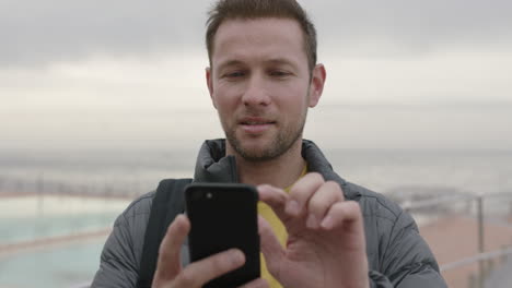 Retrato-De-Un-Hombre-Encantador-Usando-Mensajes-De-Texto-Por-Teléfono-Junto-Al-Mar-Sonriendo-Relajado