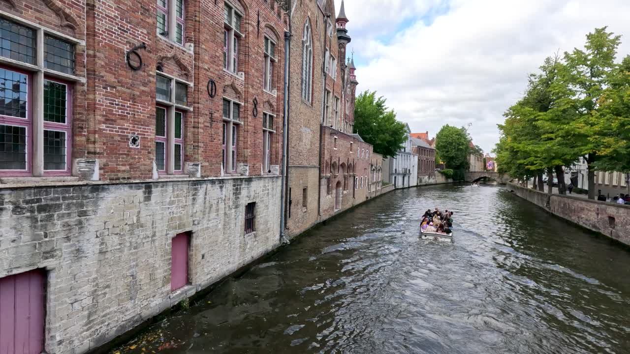 Tourist boat cruises along historic canal with brick buildings and trees on a summer day