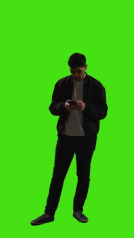video vertical de larga duración de un joven que envía mensajes de texto en un teléfono móvil de pie contra una pantalla verde 1