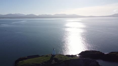 Aerial-view-across-Ynys-Llanddwyn-beacon-moving-through-shimmering-Irish-sea-at-sunrise-with-hazy-Snowdonia-mountain-range