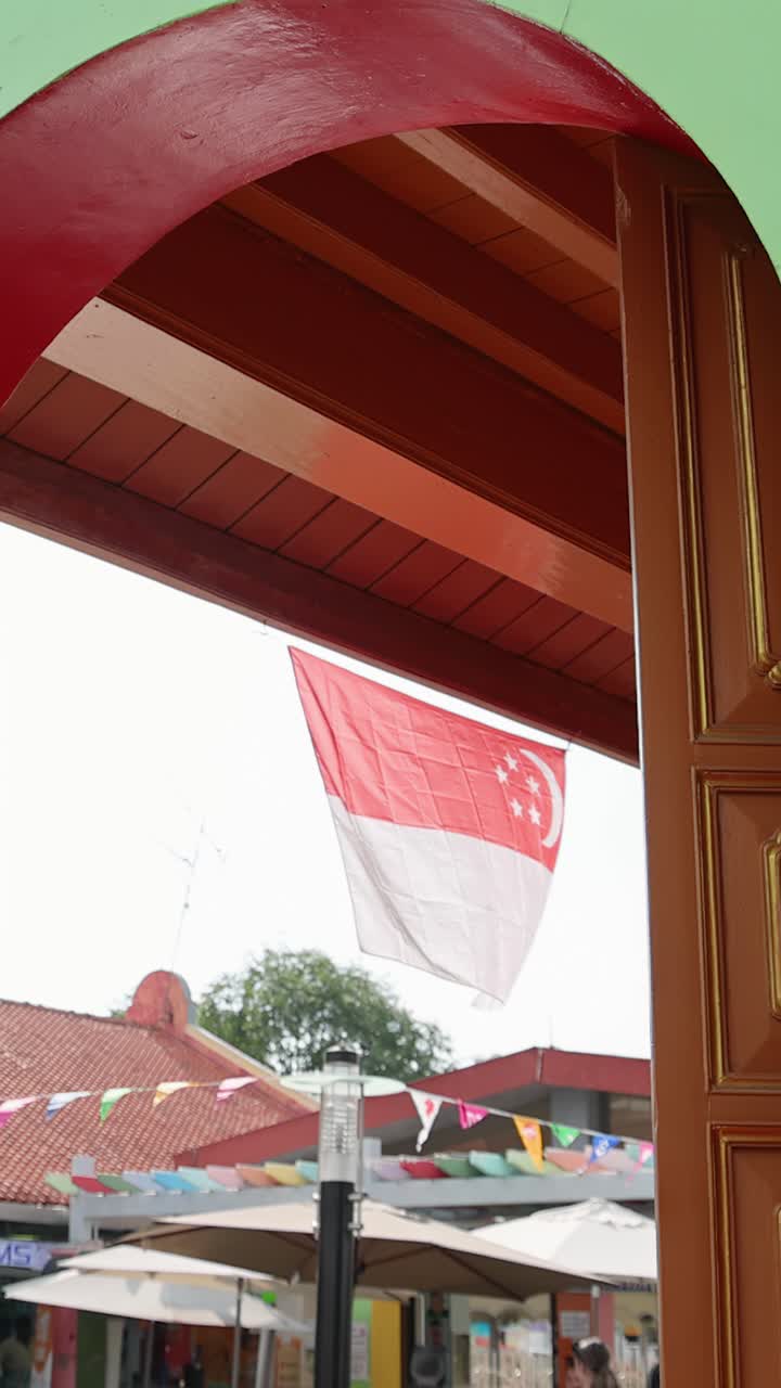 La bandiera di Singapore sventola nella brezza diurna, vista attraverso la finestra ad arco di un quartiere storico e vivace.