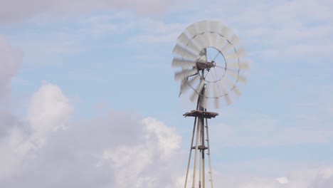 Close-up-4K-shot-of-an-old-rustic-farm-windmill-blowing-and-spinning-in-the-wind-with-a-partly-cloudy-blue-sky-in-the-background