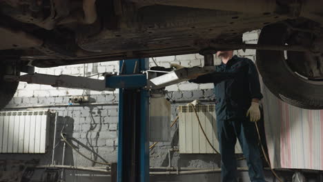 mechanic in dark uniform and gloves operating hydraulic lift in mechanical workshop, raising a car for inspection, underneath view shows vehicle suspension, tires, and metal structure