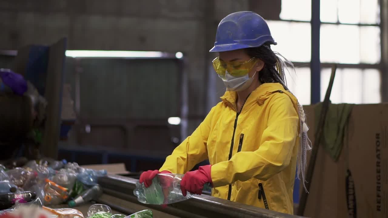 Woman-volunteer in yellow and transparent protecting glasses and mask sorting used plastic bottles at recycling plant. Separate bottles on the line, removing tops and squeeze them