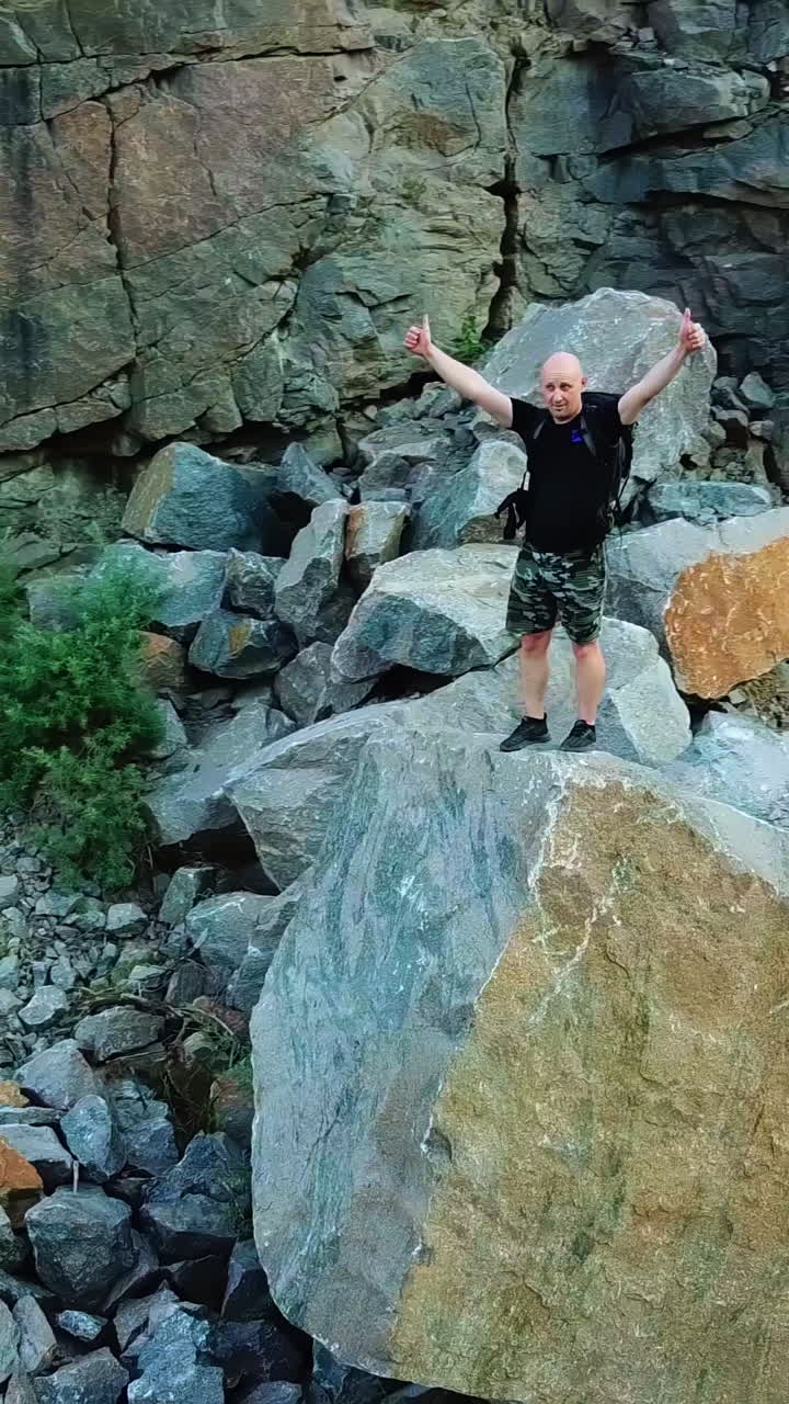 A man in a black t-shirt and a backpack comes to the edge of a large stone and raises his hands up. Tourist got to the center of stone quarry. Vertical video