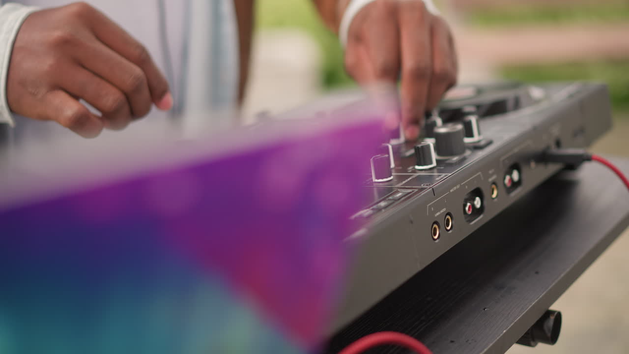 Mani di DJ di colore sul controller, set di strada diurno soulful con copertina colorata del laptop, primo piano su manopole e jog wheel, giacca testurizzata e polsino, bokeh caldo all'aperto, mixaggio ritmico e fluido.