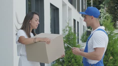 beautiful woman meets a delivery man who gives her a parcel box beside her home, courier checks information on tablet. home delivery, online shopping