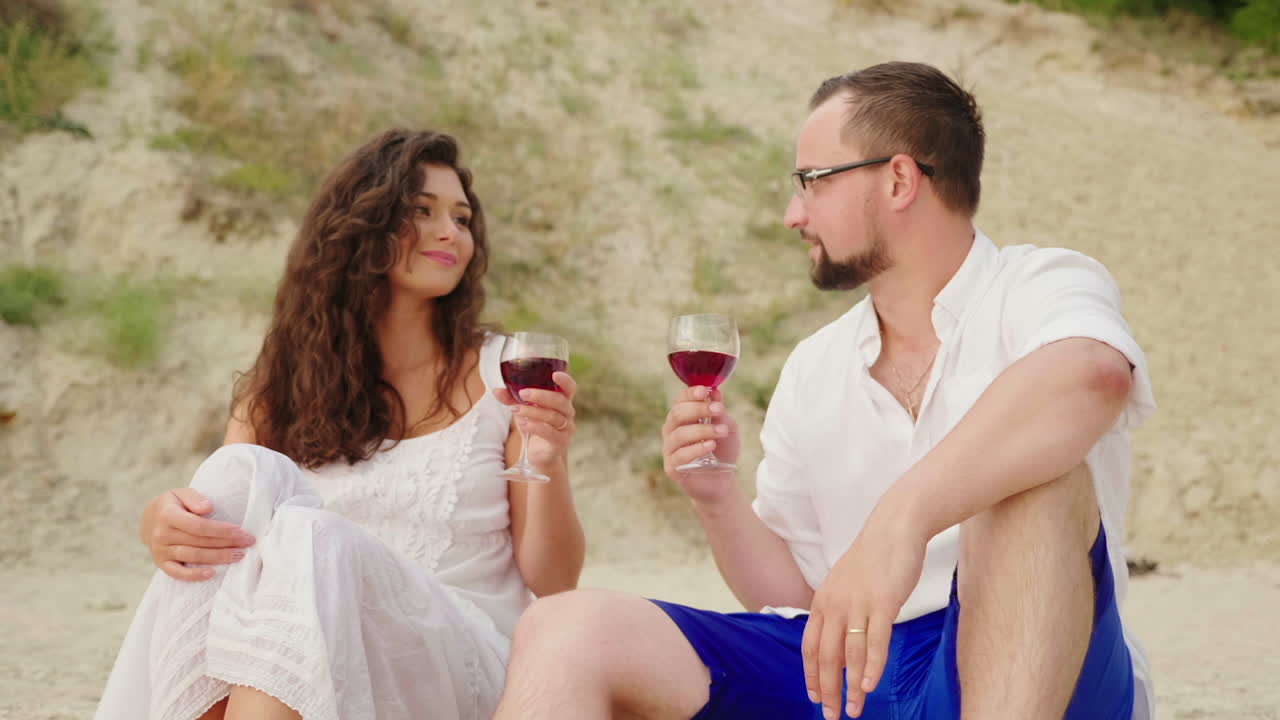 Una giovane coppia beve vino in riva al mare al tramonto. Sono seduti sulla sabbia, fanno tintinnare i calici - Anniversario
