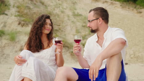 a young couple is drinking wine by the sea at sunset they sit on the sand clink glasses anniversary