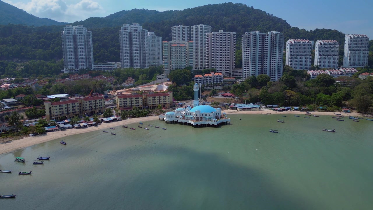 Mezquita flotante de Tanjung Bungah, Penang, Malasia, con barcos de pesca tradicionales a lo largo de la costa y edificios modernos al fondo. Espectacular vista panorámica aérea con dron.