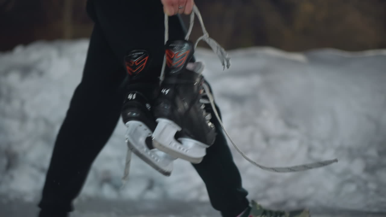 Side view of skater walking holding black ice skating boot by laces near packed snowy ice surface in cold outdoor setting, showing close-up of hand and skates with winter environment in soft blur
