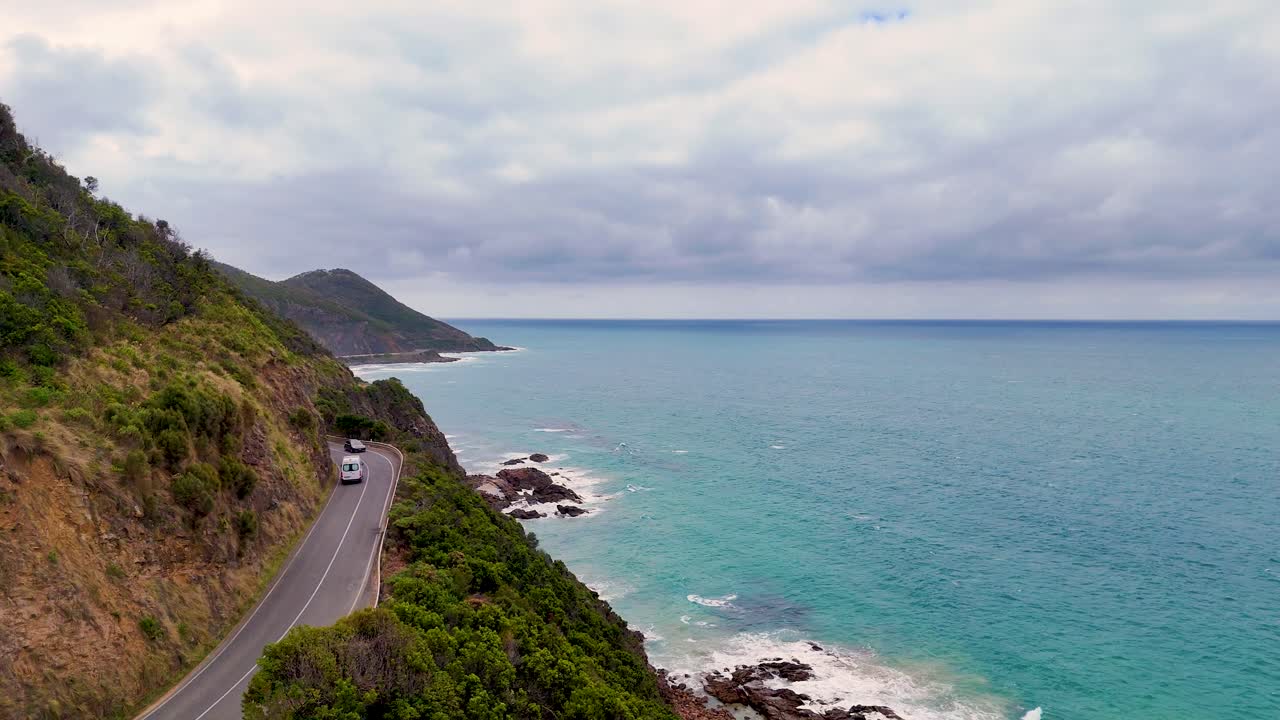 Aerial view of a car driving along the scenic Great Ocean Road, with lush greenery and turquoise ocean under a cloudy sky