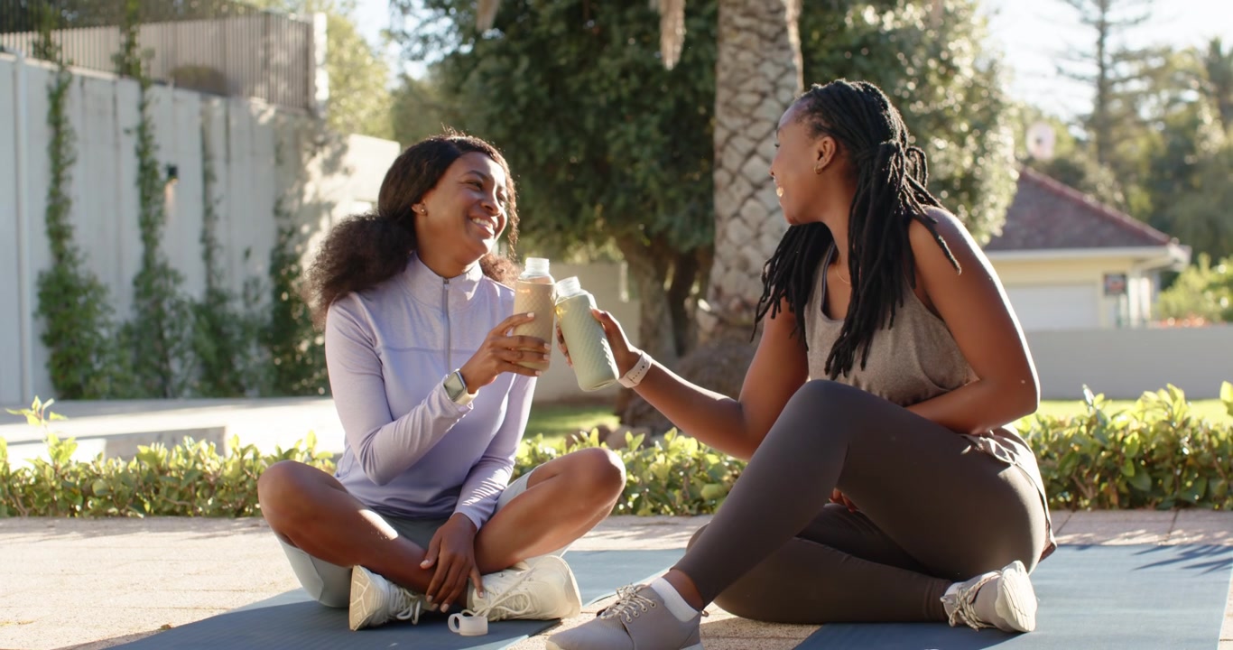 African American women friends in gear on yard mats removing cap passing bottles laughing on break