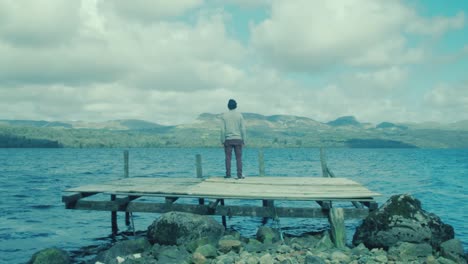 Young-man-sits-on-pier-looking-out-over-scenic-view-on-lake