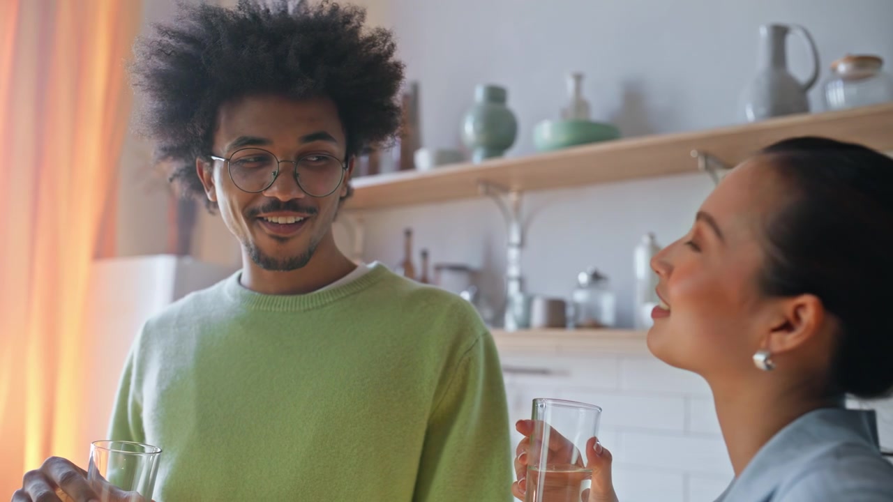 Interracial pair drinking water glasses home kitchen. Hydration wellness habit.