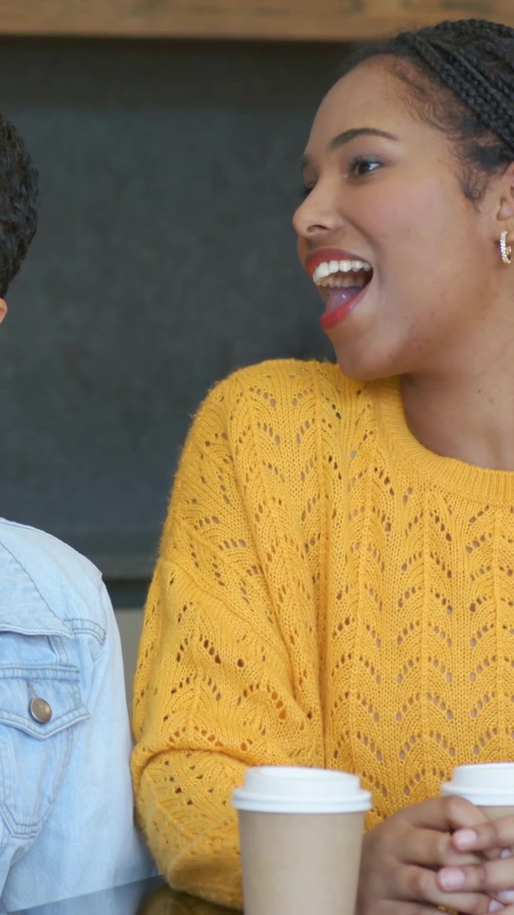 Vertical video: Laughing woman in yellow top responding to friend talking at cafe with paper cup