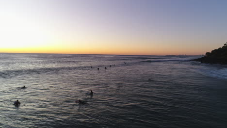 Die-Silhouette-Aus-Der-Luft-Zieht-Sich-Zurück-Und-Folgt-Einer-Gruppe-Von-Surfern,-Die-Während-Des-Sonnenaufgangs-An-Der-Goldküste-Von-Burleigh-Heads,-Queensland,-Auf-Die-Richtige-Welle-Warten