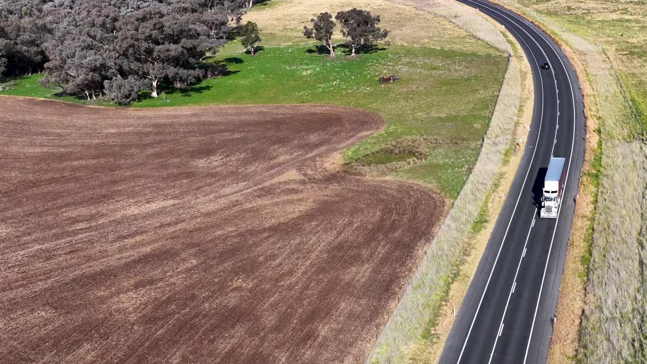 A white semi-truck travels along a winding rural highway bordered by plowed fields and sparse trees, captured in bright daylight from an aerial perspective