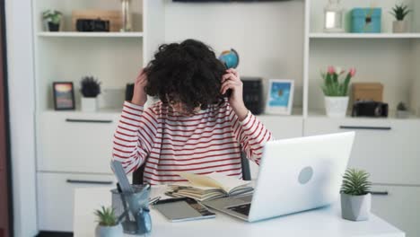 a girl with curly hair and an attractive face is listening to music in wireless headphones while sitting at a desk in a bright office and enjoying the song