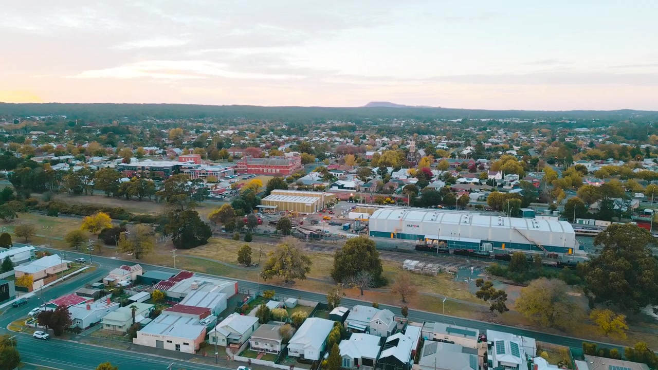Živé záběry z dronu nad Ballarat East, ukazující obytné domy, nádraží Ballarat a zářící oranžovou oblohu při východu slunce na podzim.