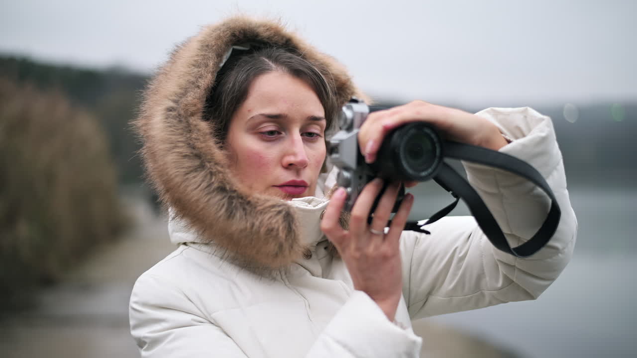 A woman stands by the water, focusing her camera on the beautiful surroundings. She is dressed warmly, capturing the serene landscape under a gray sky