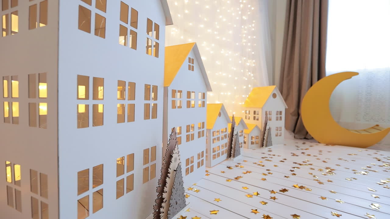 Big and small white houses of cardboard with yellow roofs. Festive lights illuminate diorama. Christmas decor in studio.