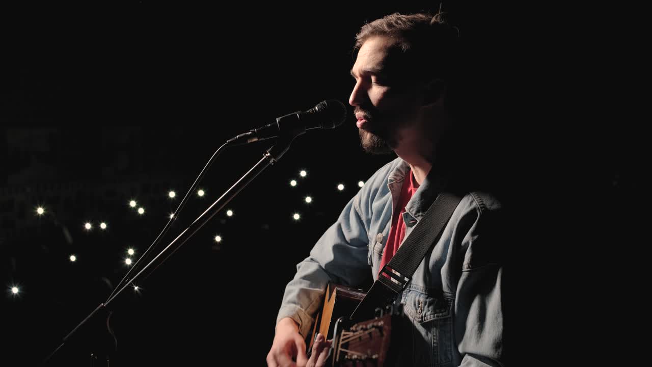 un músico está tocando la guitarra en un gran escenario con miembros de la audiencia sosteniendo linternas en el fondo. el auditorio está lleno de hermosas luces parpadeantes.