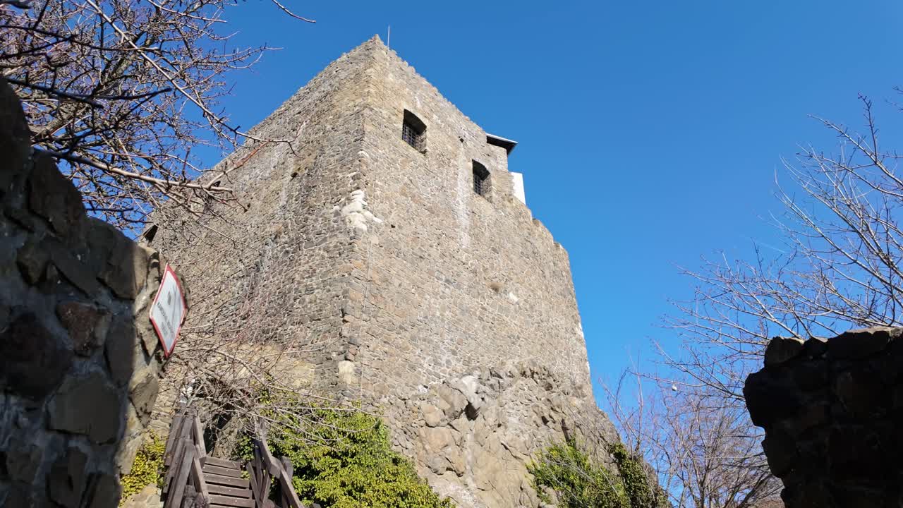 Looking up at the medieval tower of Hollókő Castle, Hungary. This stone structure stands tall against a clear blue sky, showcasing its historical significance and defensive architecture.