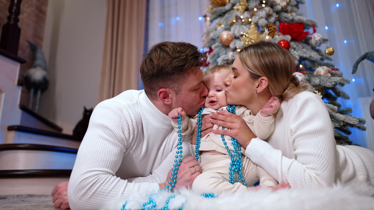 Loving parents lie on the floor with their baby in the middle. Mom and dad kiss their child to the cheeks. Christmas tree at backdrop. Low angle view.