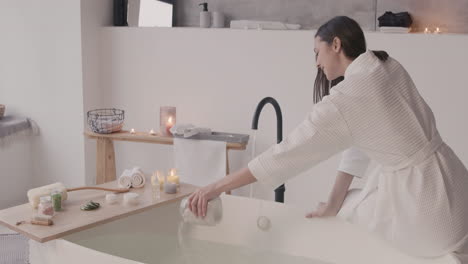 woman in bathrobe sitting on edge of bathtub while pouring bath salts on the water