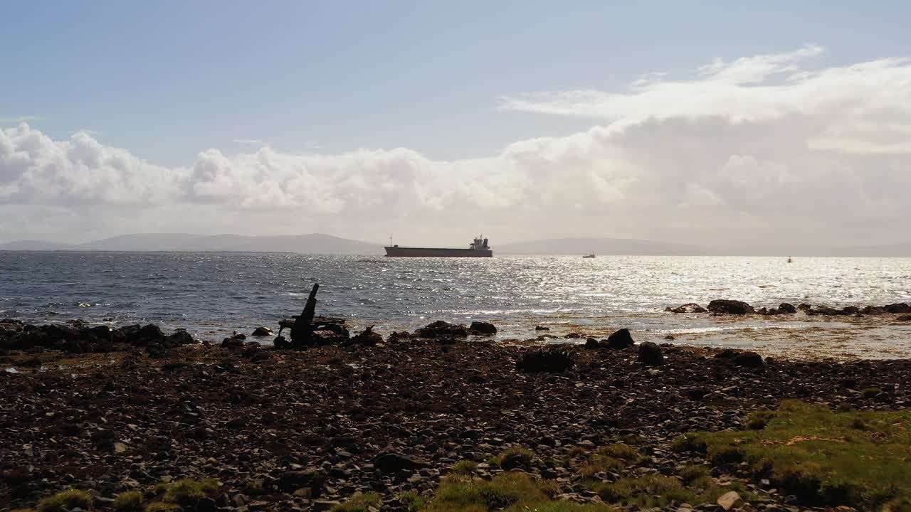 Carrello aereo sopra un'isola erbosa verso una costa rocciosa e l'oceano che si estende fino all'orizzonte con una grande nave cisterna nella Baia di Galway, Irlanda