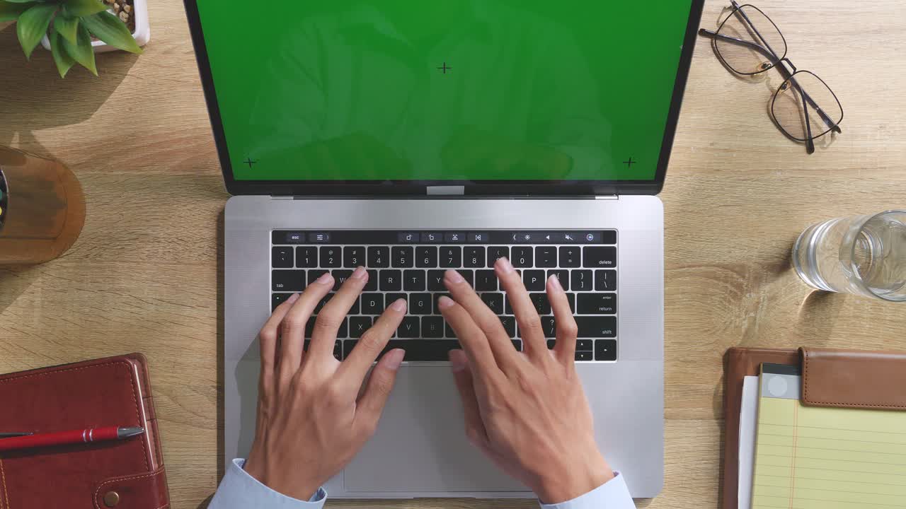 Top down view of a laptop computer with mock up green screen chromakey display on a wooden office desk next to notebook with pens, glasses, and a glass of water. Slow zoom out, close up