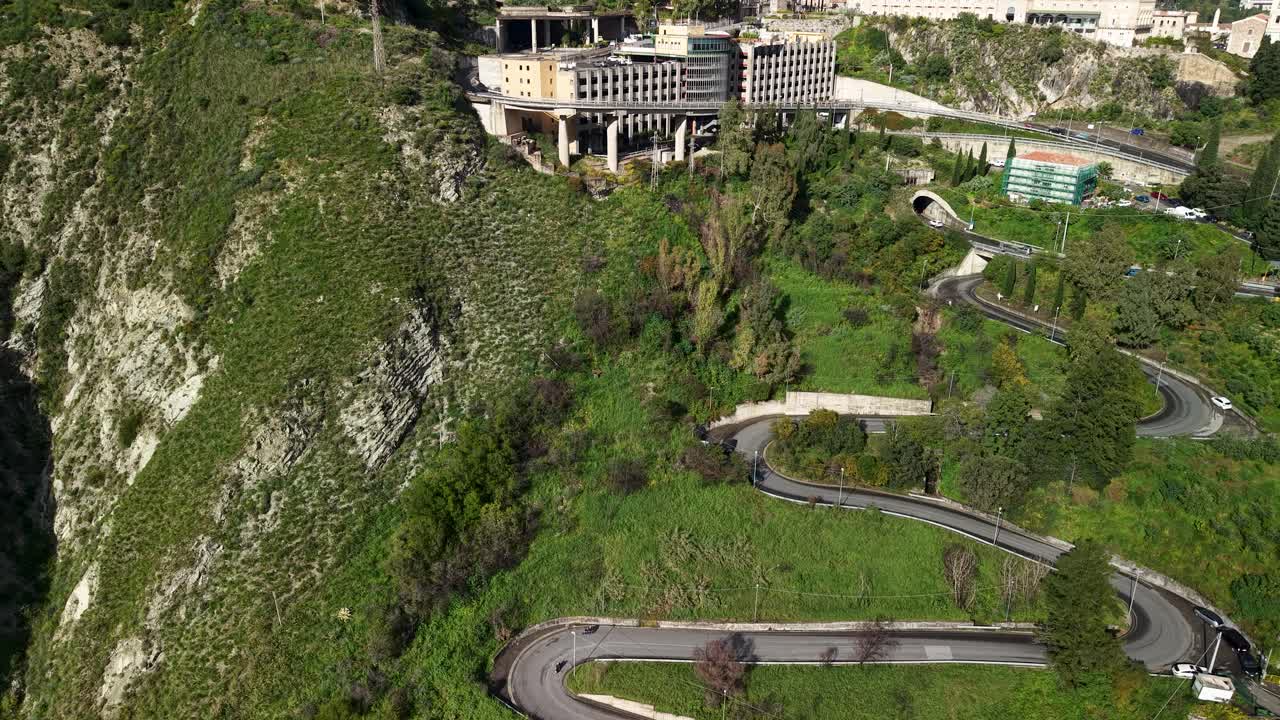Aerial view tracking winding mountain road toward Taormina showing tight curves across steep terrain