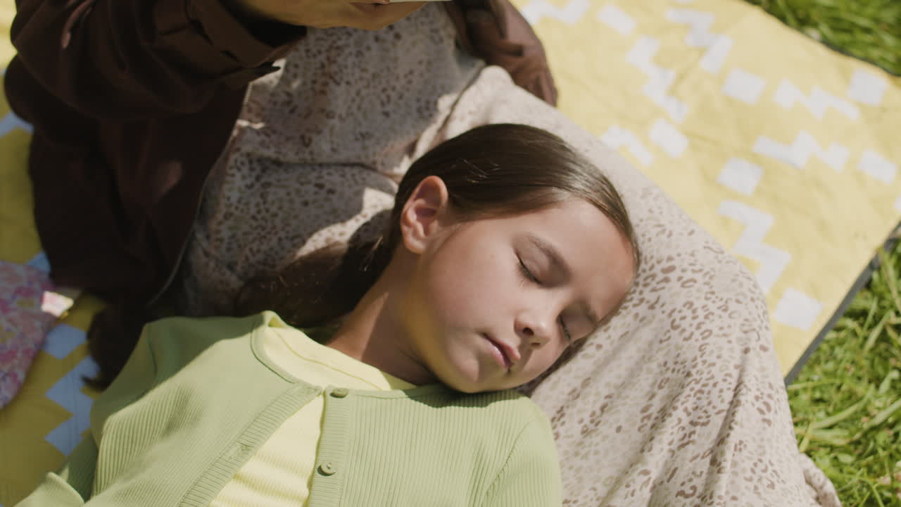 Girl sleeping on a picnic blanket while a woman reads a book
