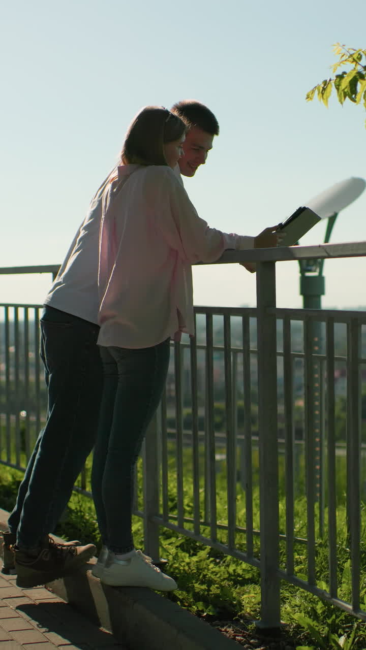 amigos apoyados en una barandilla de hierro compartiendo un momento alegre mientras el niño se ríe cubriendo la cara mientras la niña señala la tableta, el fondo presenta personas distantes borrosas contra la vegetación y la brillante luz del sol