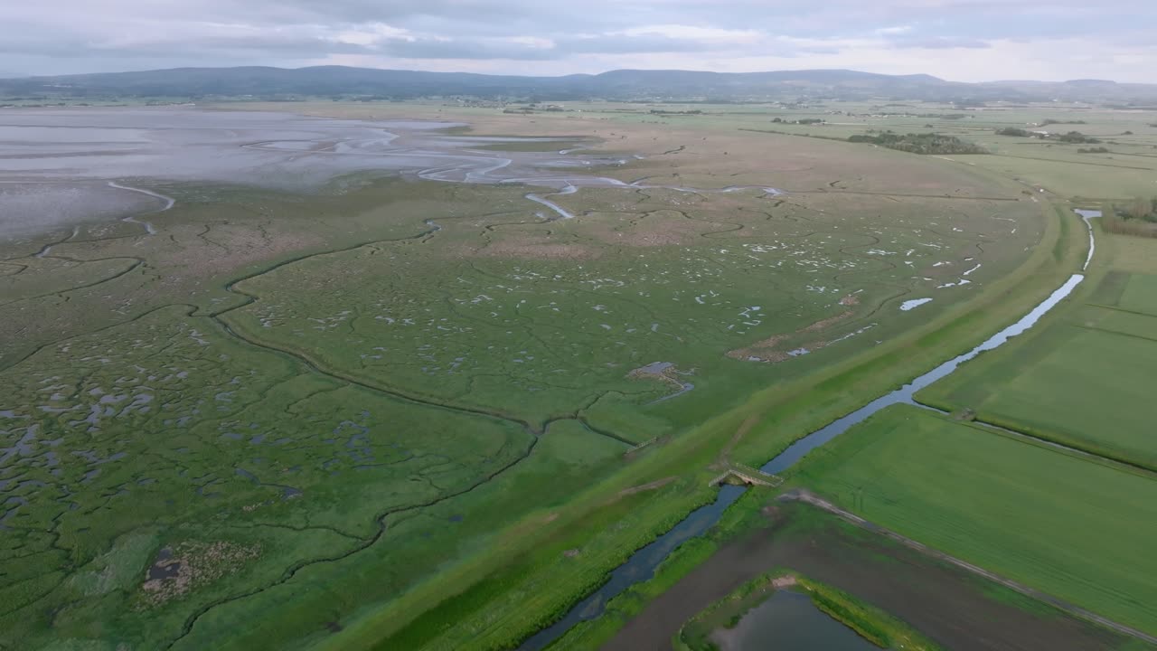 Panning aerial view of arable farmland and reveal of coastal grassland and flat tidal sands during golden hour. Pilling Sands, Lancashire, UK.