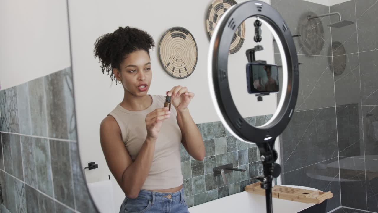 African American woman 20s filming on smartphone with ring light in bathroom, demonstrating dropper