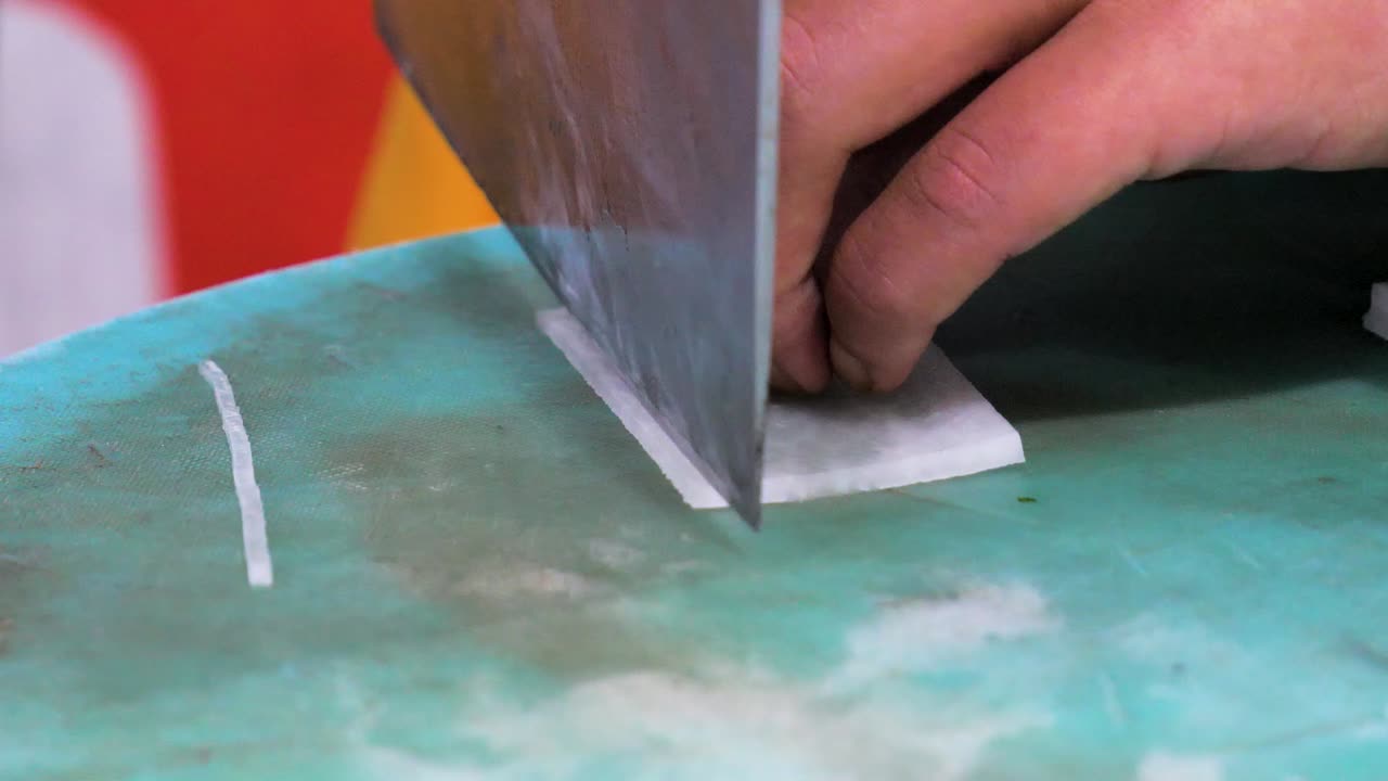 A chef demonstrates precision cutting techniques, slicing a radish with a large cleaver on a worn chopping board, showcasing traditional culinary skills in a Chinese cooking school.
