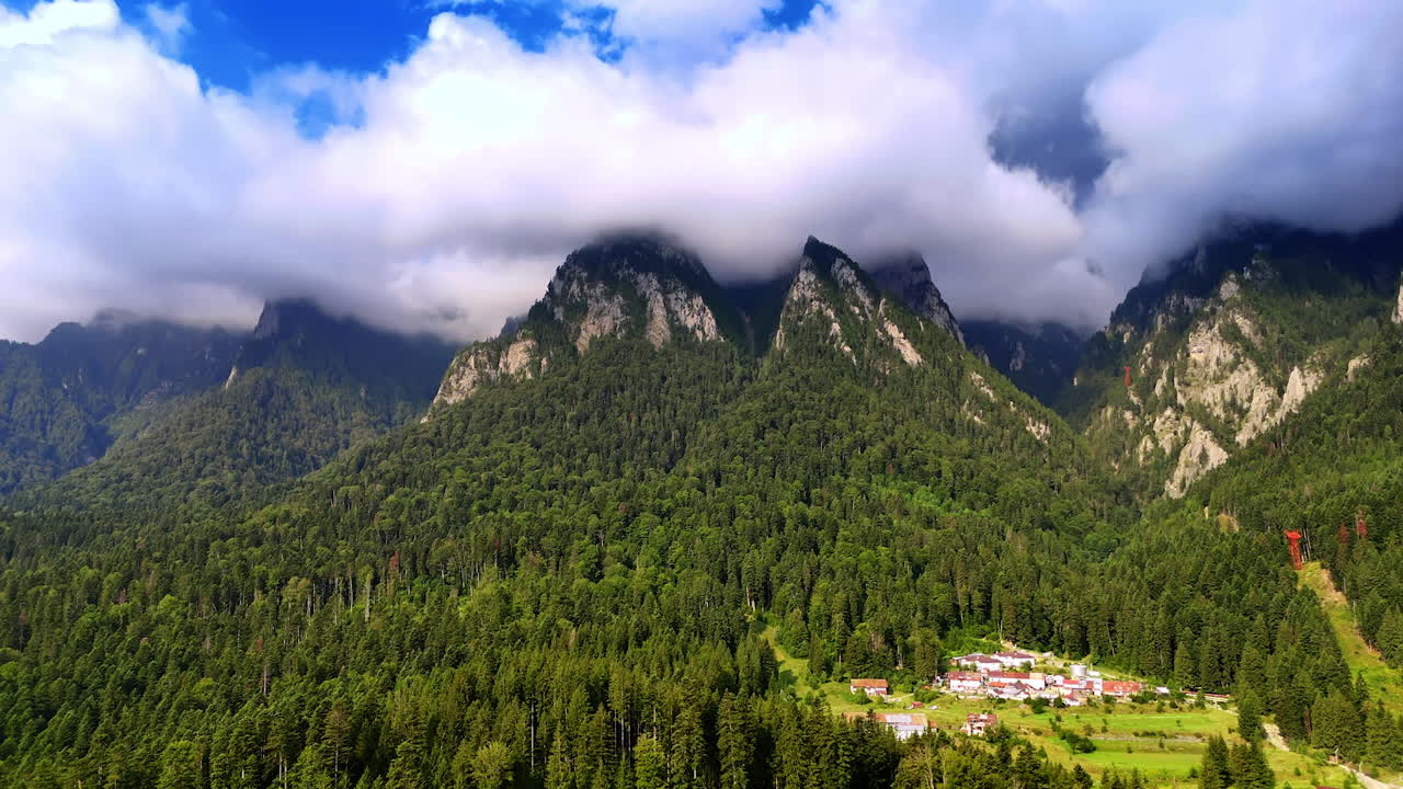 Montagne con nuvole sopra la foresta e il villaggio durante il giorno. Le nuvole coprono le cime delle montagne sopra una foresta verde e un piccolo villaggio sottostante durante il giorno.