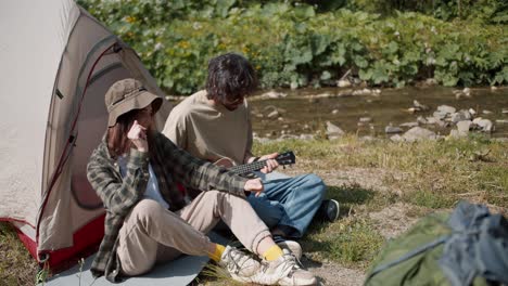 shooting near a man and a girl are sitting near a tent a man is playing a guitar a girl is listening to him and dancing they are sitting near a mountain river