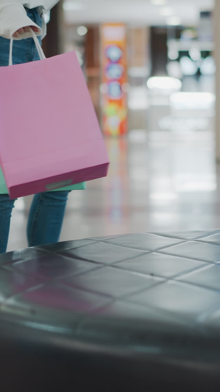 vista parcial de una dama con un traje blanco caminando hacia un asiento de cuero con bolsas de compras en la mano, colocando bolsas rosas y de menta en un asiente de cuero en un centro comercial, fondo borroso con un entorno comercial moderno