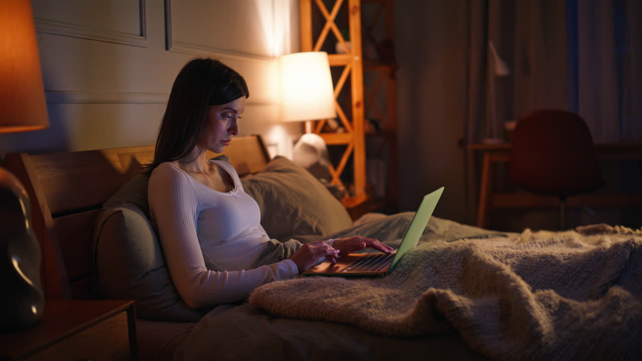 Freelance serio/a lavora di notte sdraiato/a in una camera da letto buia. Donna concentrata digita sul portatile.