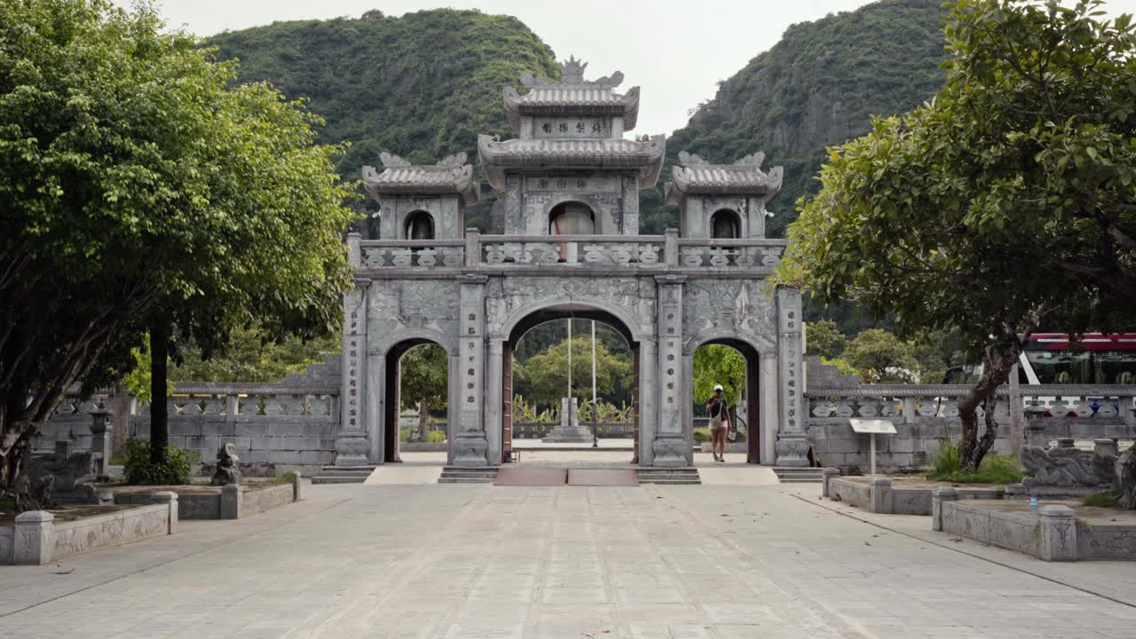 detailed view of the ancient stone gateway at Thai Vi Temple in Tam Coc, Vietnam, as a tourist enters and captures photos. Surrounded by lush trees and dramatic mountains