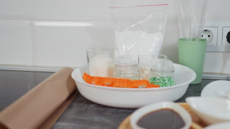 kitchen counter with wooden tray holding ceramic bowls filled with sauce, spices, and sugar, butter on plate, glass jars with ingredients, baking tools, and kitchen utensils neatly arranged