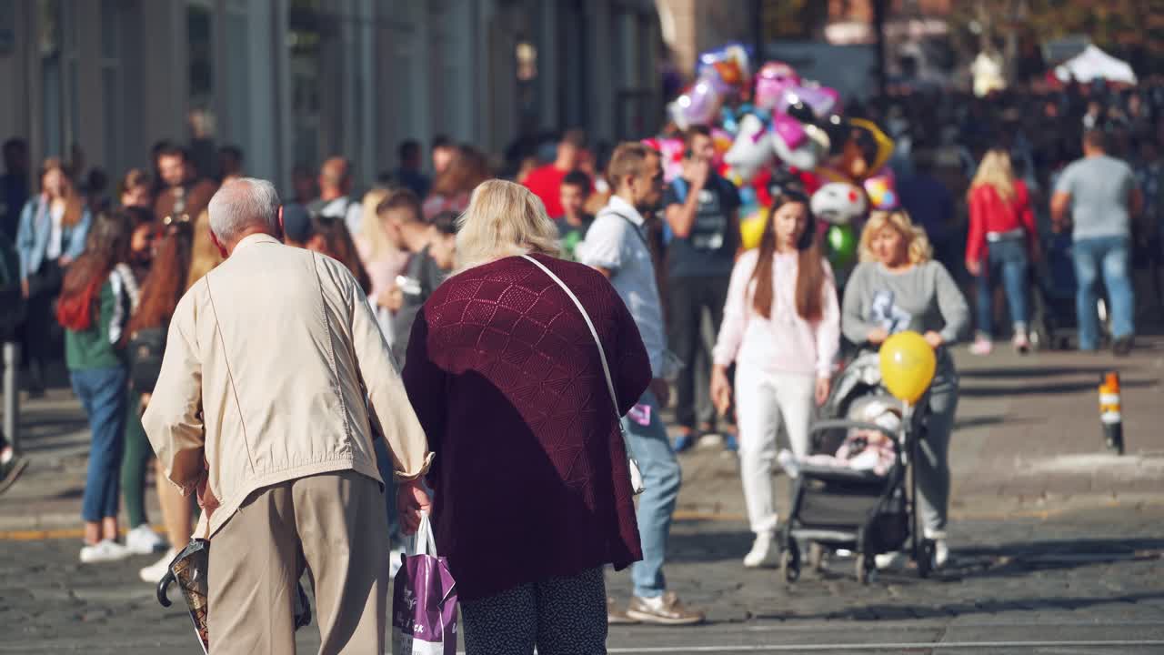 Many people in the city. Backside view of old people walking on the sidewalk on crowded city background. Colored balloons sold in the street.